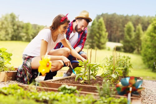 In the image, a young woman and a man are gardening together in a well-maintained outdoor space, possibly part of a front or back garden in Acton. The woman, wearing a casual white t-shirt and plaid shorts, is kneeling on the ground, planting or tending to small green plants within a raised wooden bed. She has a pink headband and sunglasses, indicating warm weather. The man, dressed in a red and blue checkered shirt and a straw hat, is also kneeling nearby, assisting with the garden work, holding a gardening tool. Surrounding the pair are various garden elements including the rich, dark soil in the planting bed, a nearby lawn with fine grass, and a blurred background of trees and shrubs, suggesting a lush, natural environment. The garden appears to be in the midst of active cultivation, with seedlings or young plants visible and garden tools minimally present in the scene. The weather is sunny and clear, providing bright natural light that highlights the greenery and casual outdoor atmosphere typical of a well-kept garden in the Acton area, reinforcing the relevance of professional gardening services offered by Gardening Services Acton to maintain such outdoor spaces.